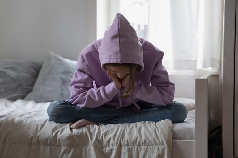 Unhappy depressed teenage girl wearing sweatshirt sitting on bed alone, feeling lonely and misunderstood, stressed sad teenager thinking about troubles, hiding face, child and psychological problem; Shutterstock ID 2170756605; purchase_order: aljazeera ; job: ; client: ; other: